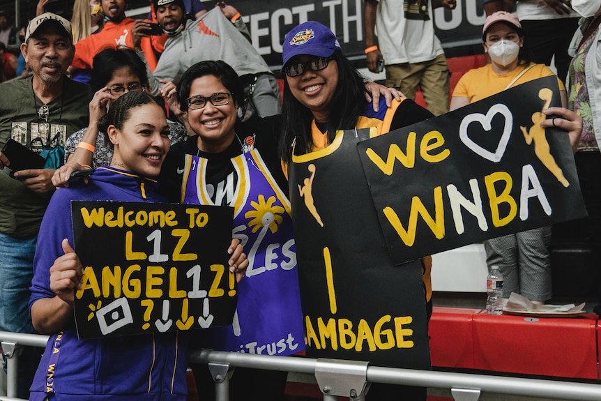 A smiling Aussie basketballer holds a sign saying "Welcome L1Z Angel1Z!" as she stands next to fans holding signs.