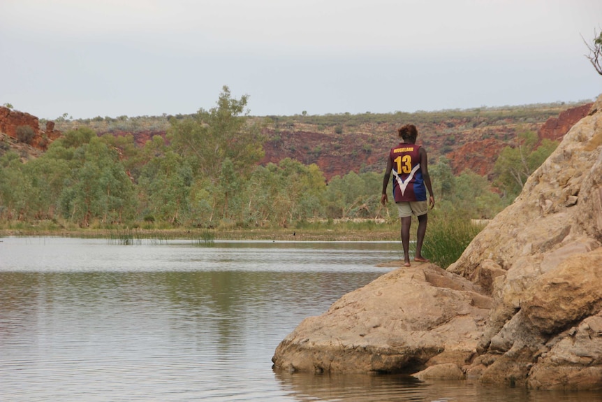 A young person with their back turned walking alongside a rocky river bed