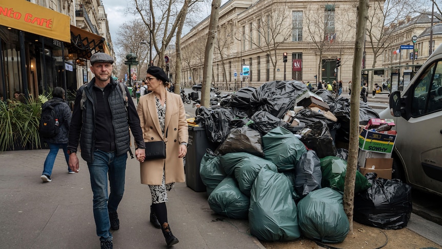 A man and woman walk past bags of rubbish piled up on the street.