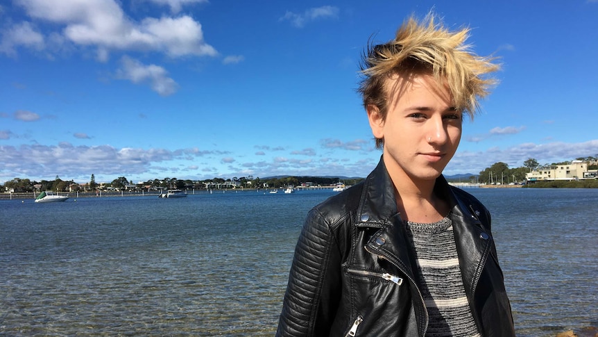 Elijah Mortlock stands by the water in Merimbula, looking out to sea. He's wearing a striped t shirt and leather jacket.