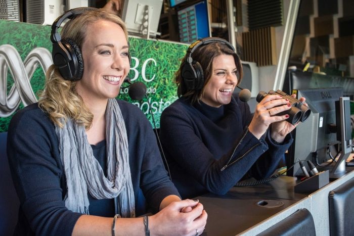 Two female broadcasters wearing headphones call an AFL game from a comment box.