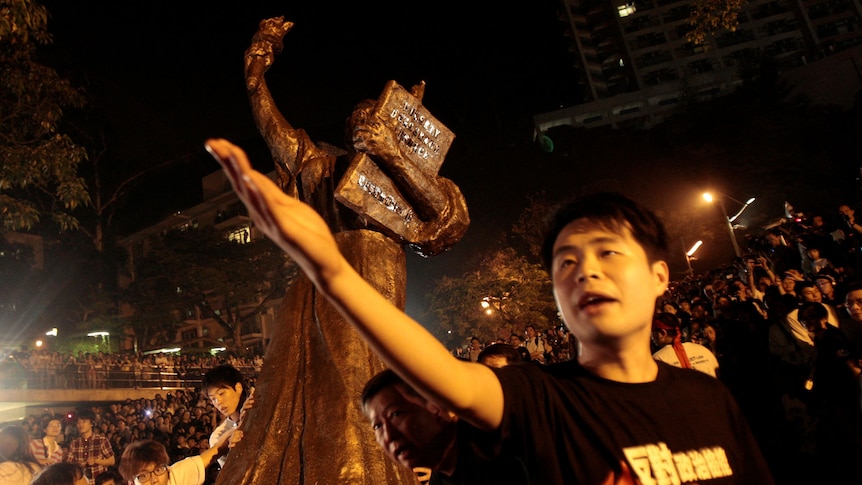A man gestures, while standing in front of a bronze statue of a woman that is being transported