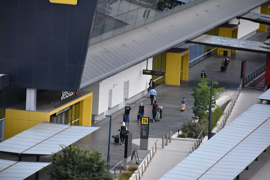 Travellers wearing masks wheel their suitcases along the front of a terminal building at Melbourne Airport.