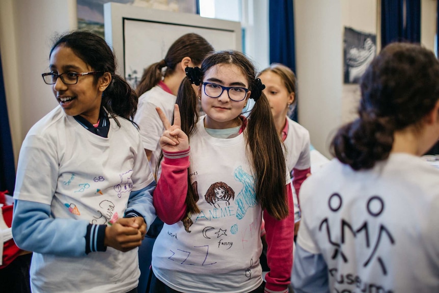 Girls wearing white shirts with Texta drawing on them, over their school uniforms, smile. One holds up hand in peace sign.