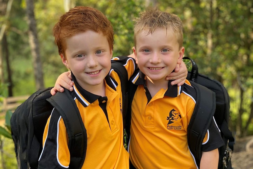 Five-year-old twins in their uniforms and back packs with their arms around eachother.