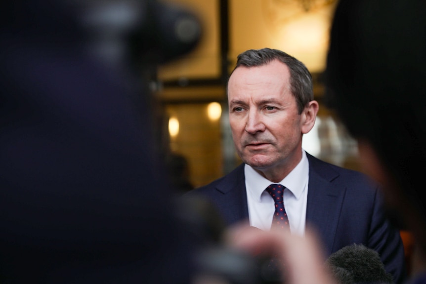 A man in a suit speaks to reporters shot from between shoulders. 