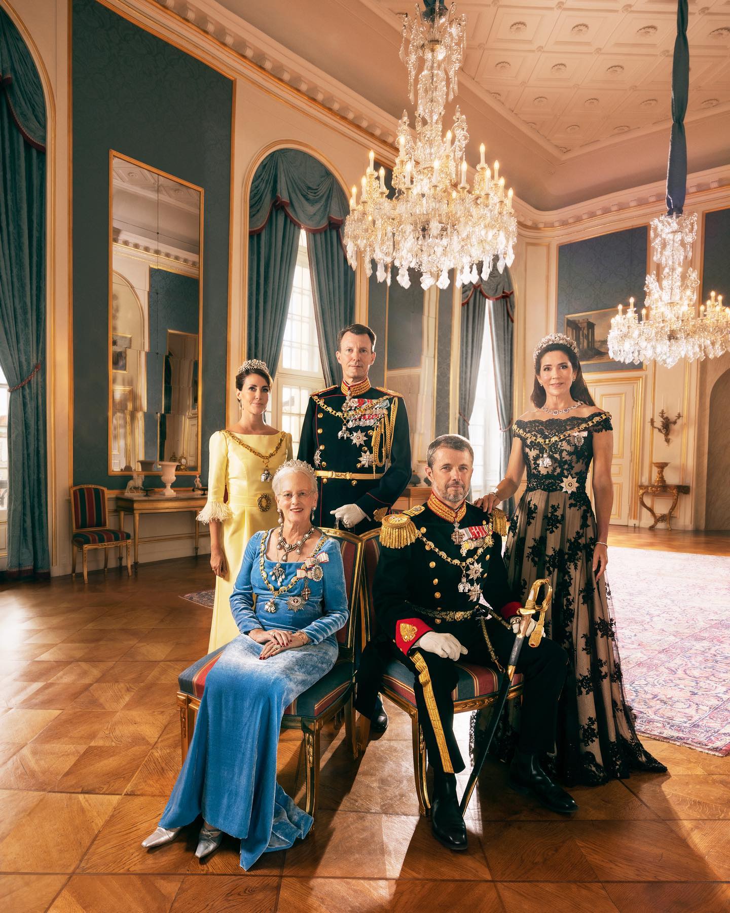 Five people in royal attire pose for a portrait, Queen Margrethe and Prince Frederik are seated at front adorned in jewels