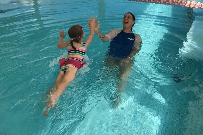 Harmony in the pool leaning back hi-fiving a little girl who is swimming towards her.