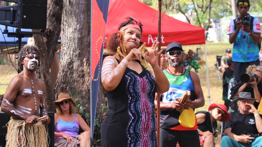 Woman dressed in bright colours and Indigenous paint makes a sign with her hands.