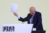 An older man wearing a blue suit holds up a piece of paper while speaking at a lectern