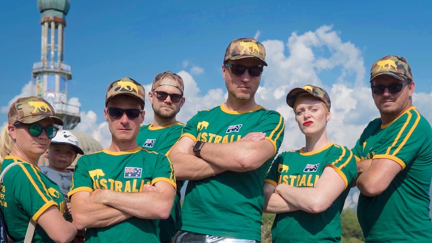 Six adults wearing green and gold sporting uniforms pose with arms crossed.
