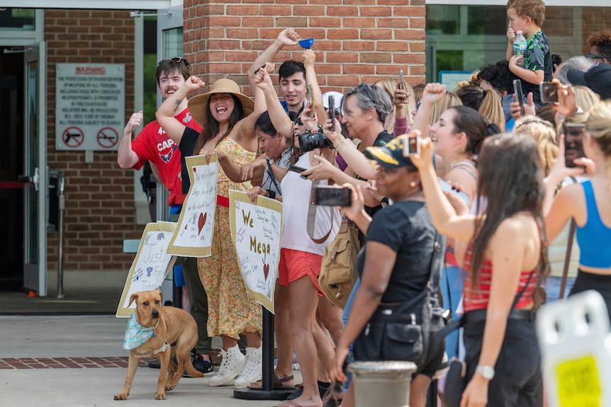 smiling people holding placards and raising their fists in the air stand outside a brick building