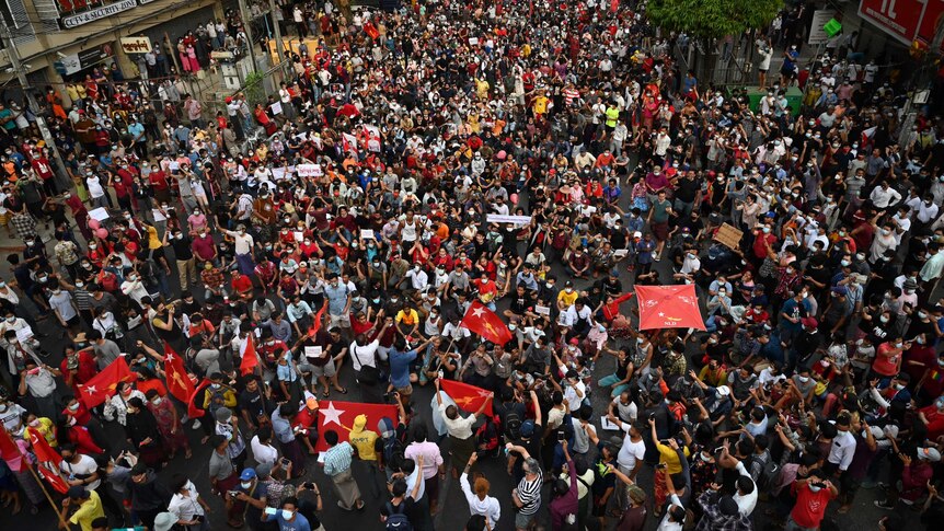 An aerial shot shows a large crowd of protesters, many wearing face masks and holding red flags with a white star.
