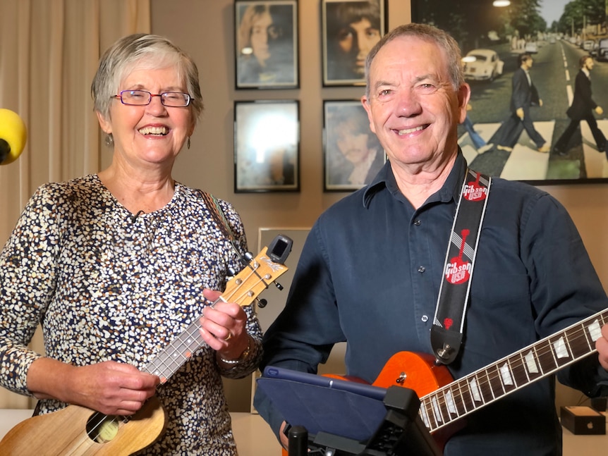 A woman with a floral print t-shirt and grey hair holds a ukulele and a man with a navy shirt holds a guitar