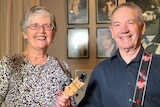 A woman with a floral print t-shirt and grey hair holds a ukulele and a man with a navy shirt holds a guitar