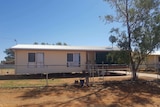 A photo of a house with stairs and a verandah in an outback setting with a large tree at the front in the fenced yard.