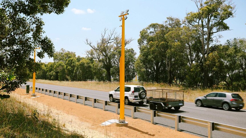 Two cars on a highway, one towing a trailer, pass by a pole mounted speed camera.