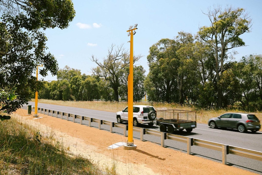 Two cars on a highway, one towing a trailer, pass by a pole mounted speed camera.