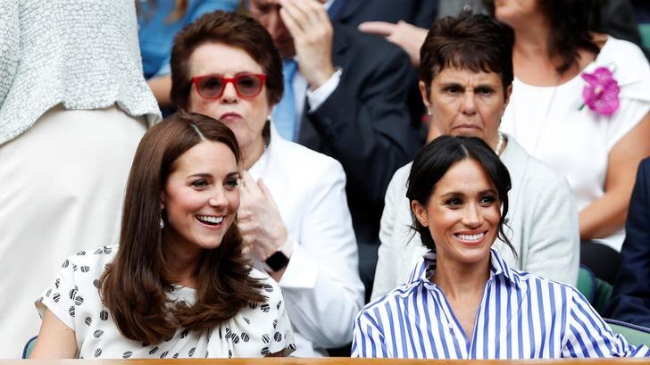 Kate sits to Meghan's left in white patterned dress, with Meghan wearing a blue and white striped shirt. Both are smiling.