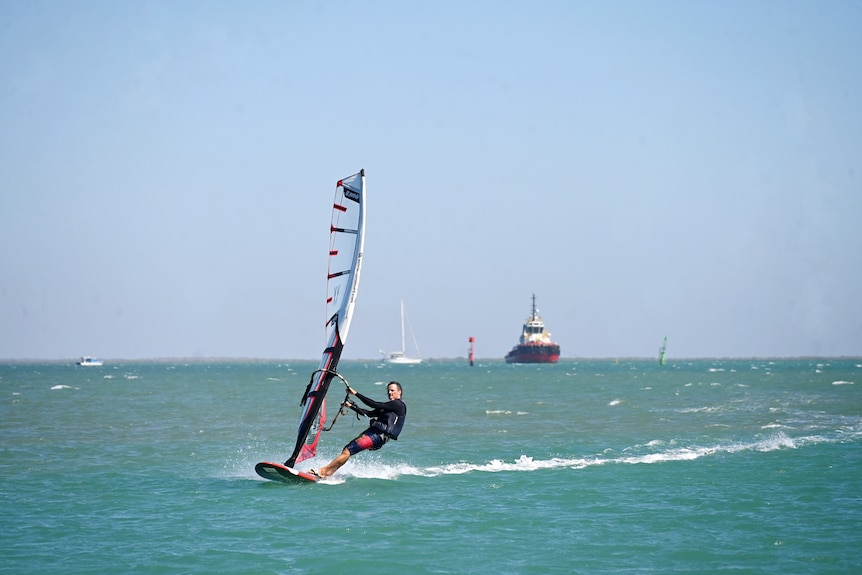 A man in a black wetsuit on a red windsurfing board and sail leaves a wake of white water with a tugboat in the background