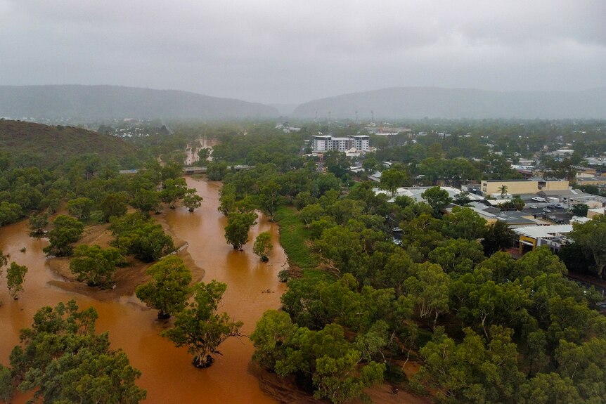 A picture taken from a drone of a flooded Todd River to the left of frame and Alice Springs on the right, with an overcast sky