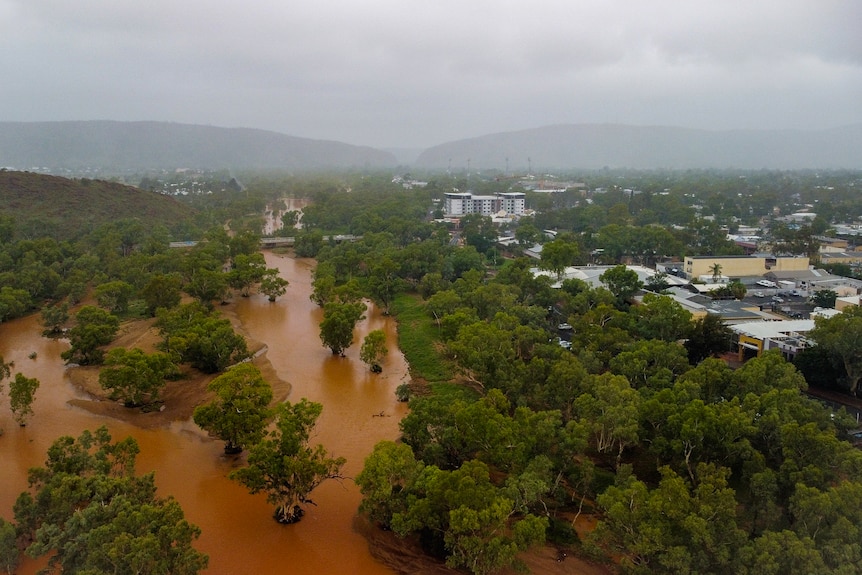 A picture taken from a drone of a flooded Todd River to the left of frame and Alice Springs on the right, with an overcast sky