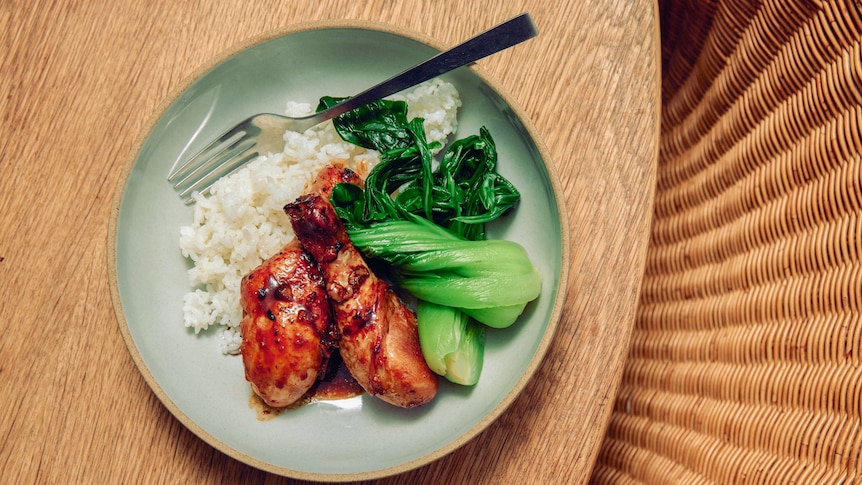 Overhead shot of a plate of honey soy chicken drumsticks alongside steamed bok choy, white rice and a fork, on a timber table.