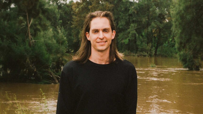 De Zilva, long-haired and in his mid-twenties, stands in front of a forest and river scene, smiling and wearing a black jumper.