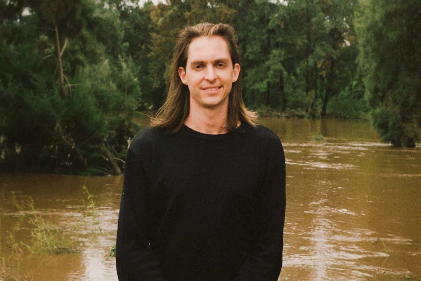 De Zilva, long-haired and in his mid-twenties, stands in front of a forest and river scene, smiling and wearing a black jumper.