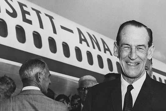 Black and white photo of a widely smiling man in a suit and tie, standing before an Ansett aeroplane and crowd of people.