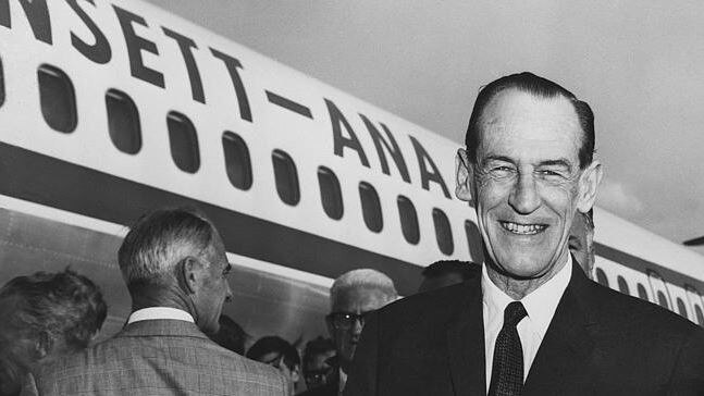 Black and white photo of a widely smiling man in a suit and tie, standing before an Ansett aeroplane and crowd of people.