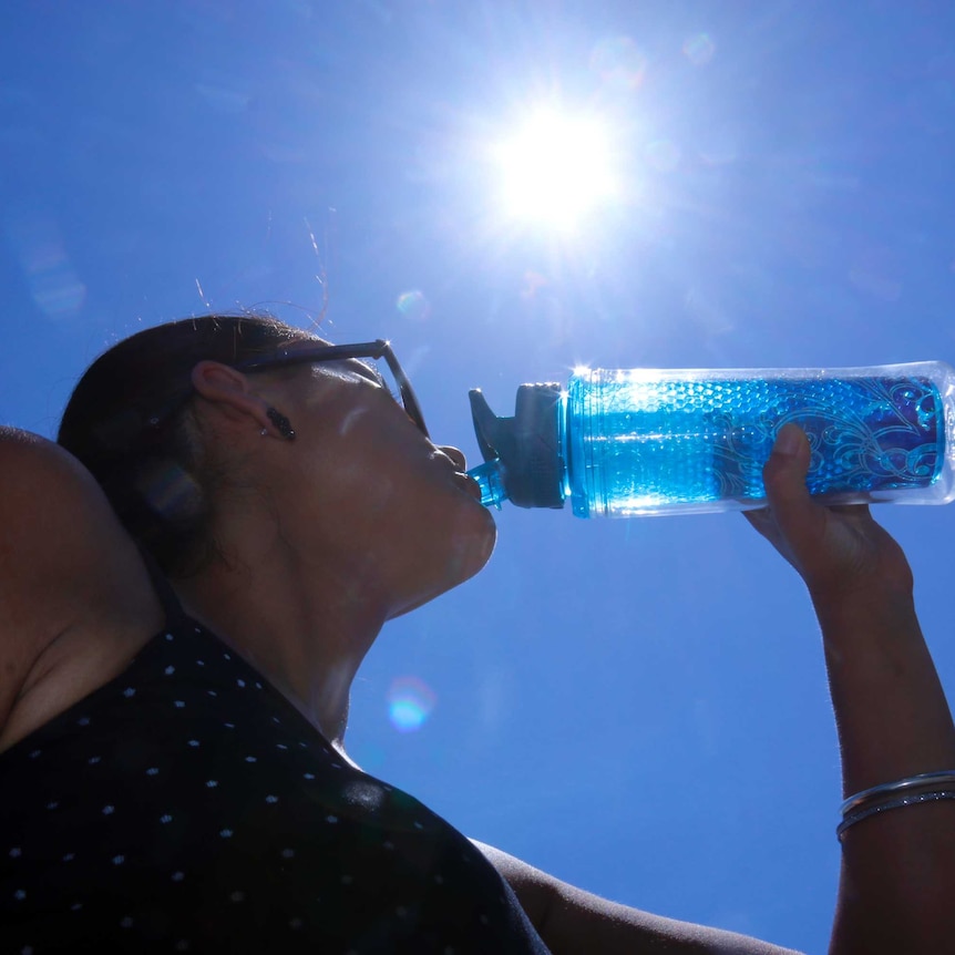 Woman drinks from her water bottle looking up towards sun in blue sky.