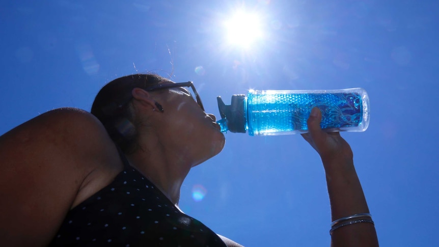 Woman drinks from her water bottle looking up towards sun in blue sky.