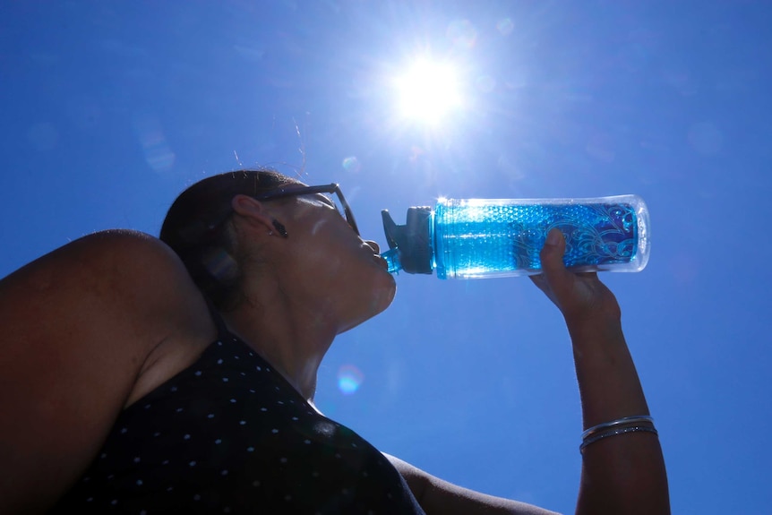 Woman drinks from her water bottle looking up towards sun in blue sky.