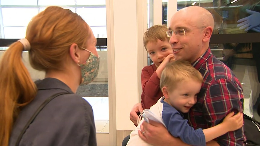 A man hugging two young boys while a woman watches on