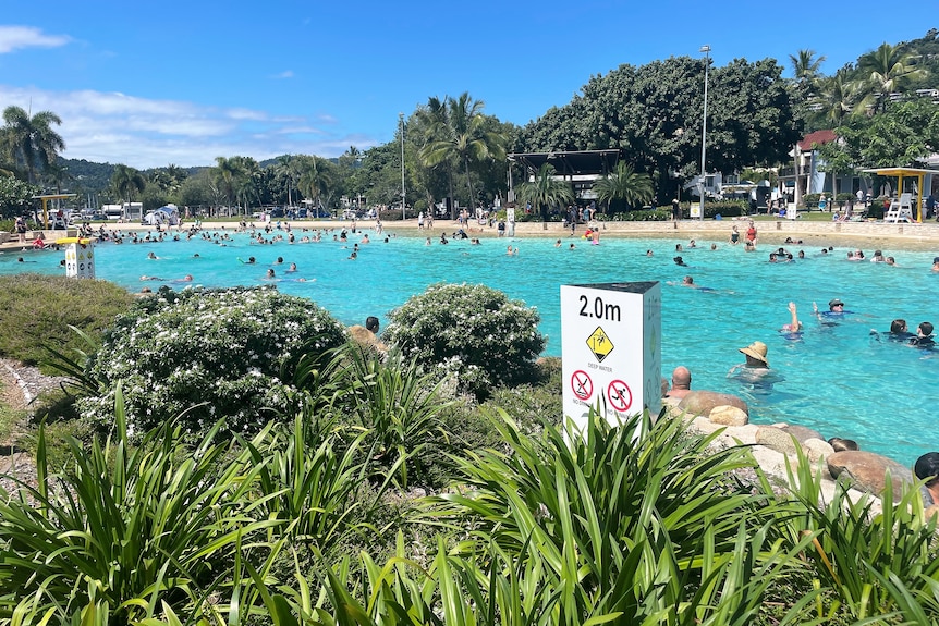 A man-made swimming lagoon with dozens of people in it.  A depth sign of 2-metres is in the foreground.