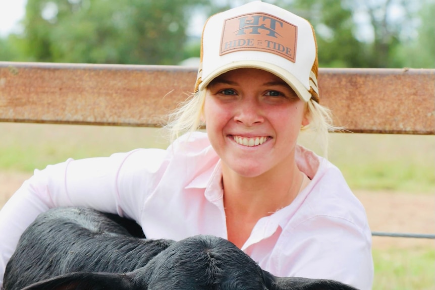 A young women is grinning while cuddling an all black calf. She's wearing a cap and a light pink button up in front of a fence