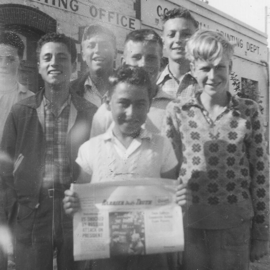 A black and white photo of a group of boys standing in front of a building with one holding a newspaper