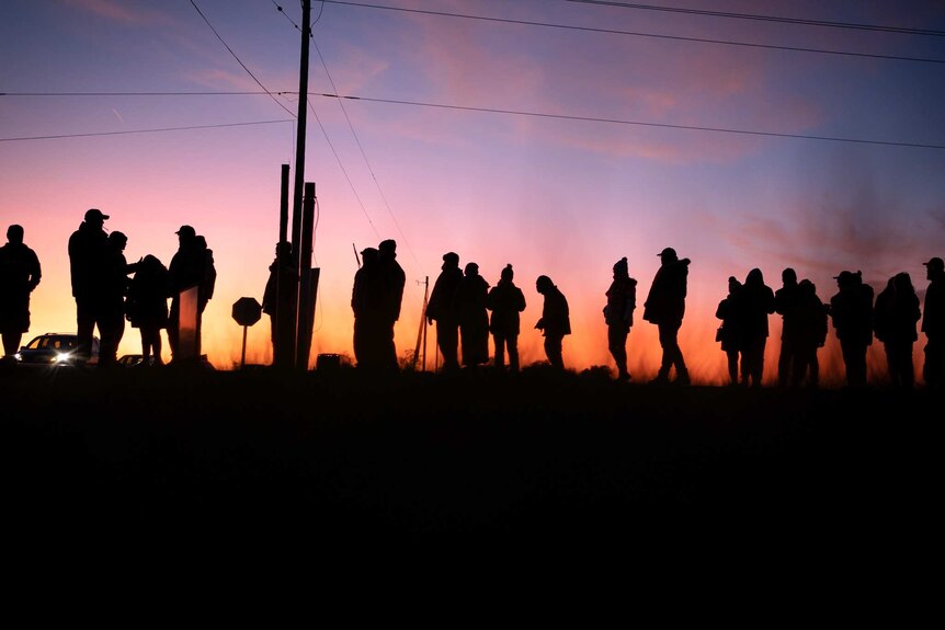 A group of people stand in a line as the sun sets in front of them