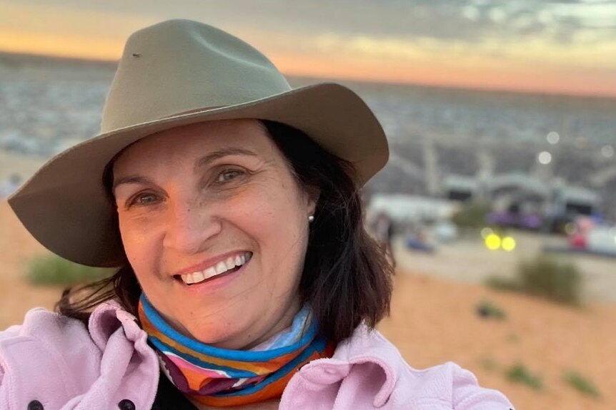 A woman wearing  an Akubra hat smiles in a selfie shot with a sunrise over the beach behind.