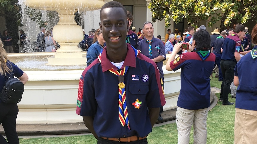 A young South Sudanese man stands on a lawn dressed in a Scout uniform.