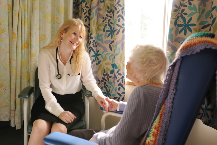 A young woman with a stethoscope around her neck holds the hand of an elderly patient as they sit together at a care facility.