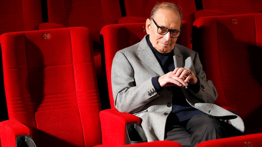 A man in a jacket sits in a theatre with red seats.