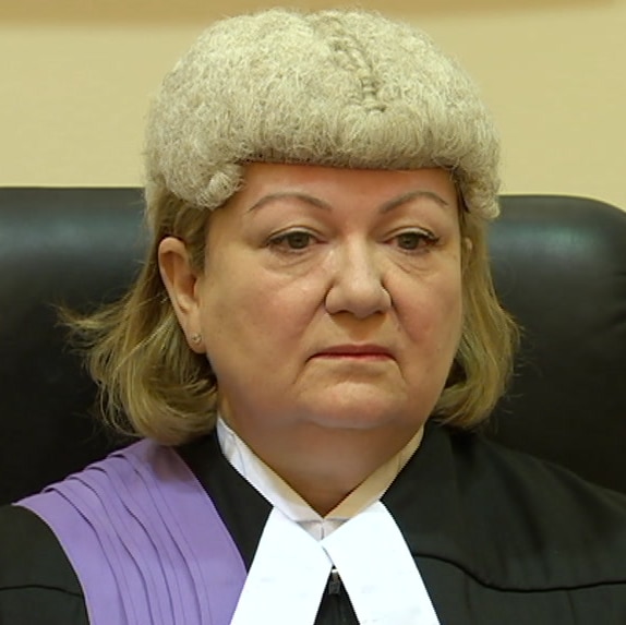 A woman with blonde hair wearing a judge's wig looks stern in a black chair