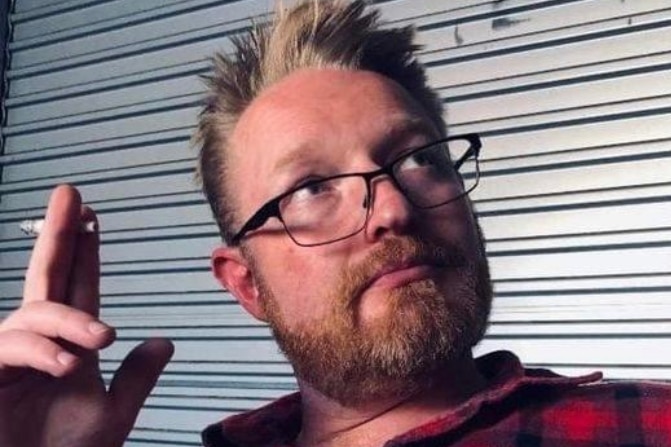 A young man with red hair and black thin framed glasses looks up, holding a cigarette.