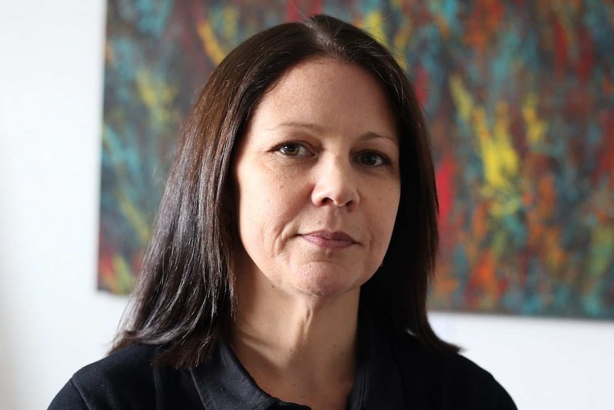 Karinda Taylor looks thoughtfully towards the camera as she stands in front of an Aboriginal artwork in an office.