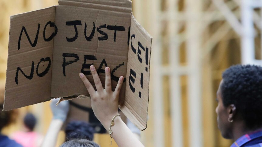 A woman holds a cardboard placard with 'No justice, no peace' sign on it.