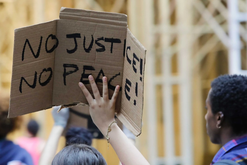 A woman holds a cardboard placard with 'No justice, no peace' sign on it.