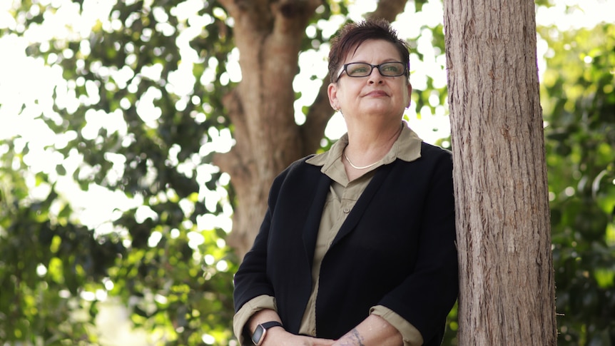 A woman with short hair and glasses looking up and off-camera, while leaning against a tree outside on a sunny day.