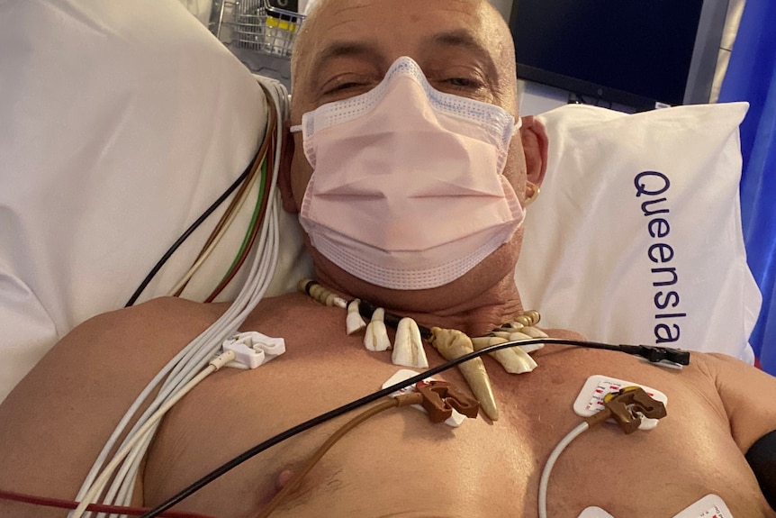 A masked man lies shirtless in a hospital bed with many monitoring devices attached to his chest.